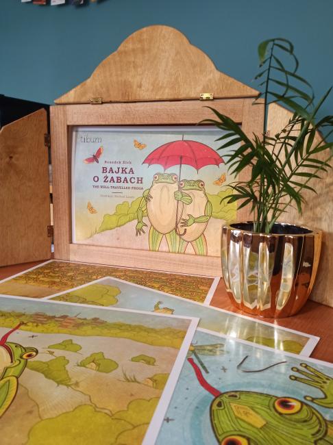 [Foto: drewniana skrzyneczka zwana "butai", w okienku znajduje się karta tytułowa "Bajki o żabach": na ilustracji dwie żaby stoją w objęciu pod czerwonym parasolem, wokół nich latają motyle.]